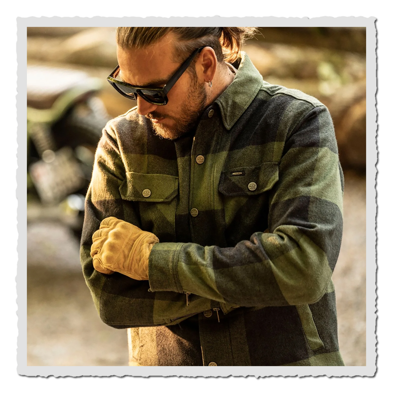 Person mit Sonnenbrille im grün-schwarzen Karo-Shirt, Oberkörper-Nahaufnahme, Outdoor, Motorrad unscharf im Hintergrund.